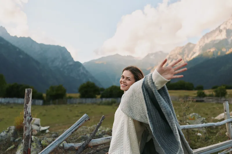 Vibrant woman outdoors, smiling while wrapped in a cozy blanket, mountains in the background, sunlit countryside scene, joyful mood and natural colors and soft breeze