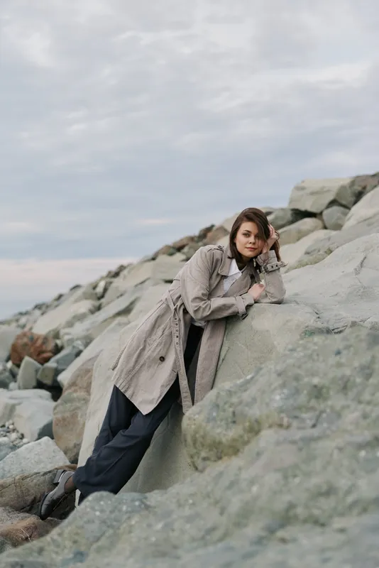 Young woman in cozy coat leans on rocky shore under cloudy sky, posing thoughtfully. Concept of autumn fashion, nature, relaxation, and casual outdoor lifestyle by the sea.