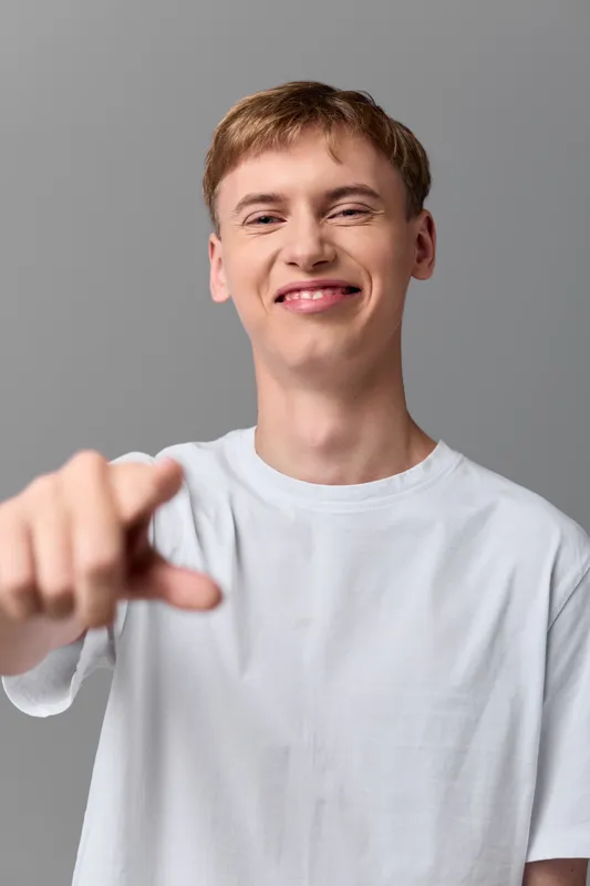 Smiling man in a white t shirt points toward the camera in a casual studio setting, presenting a friendly, confident gesture ideal for engagement, advertising, or onboarding visuals.