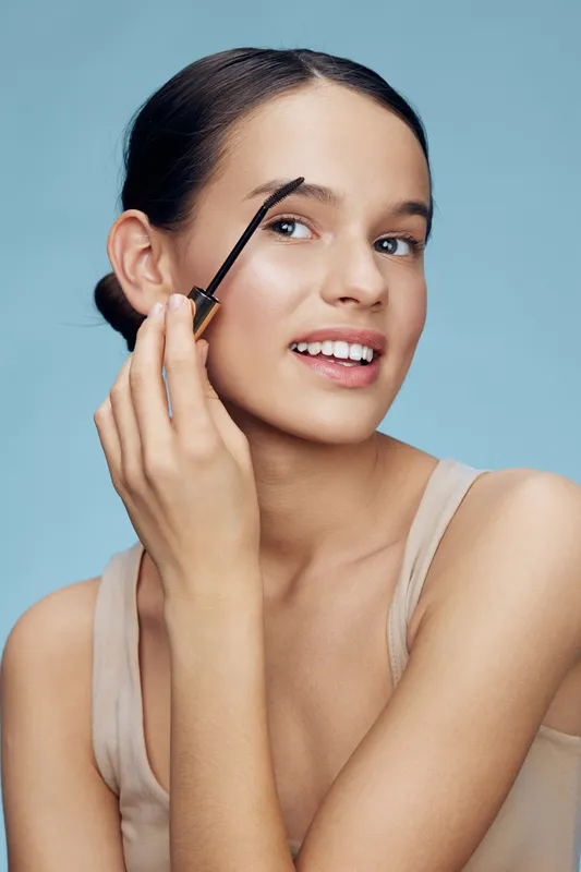 Young model applying eyebrow gel on a light blue background, showcasing beauty and skincare concepts, radiating confidence and joy with natural makeup