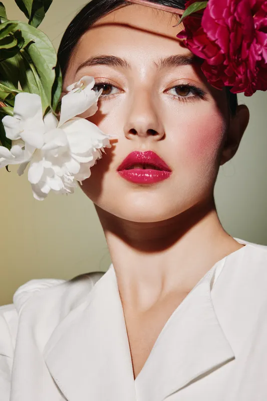Beauty portrait of a woman with creative colorful makeup featuring glossy pink lips and pink blush. She poses with large white and purplish flowers near her face. Fashion concept.