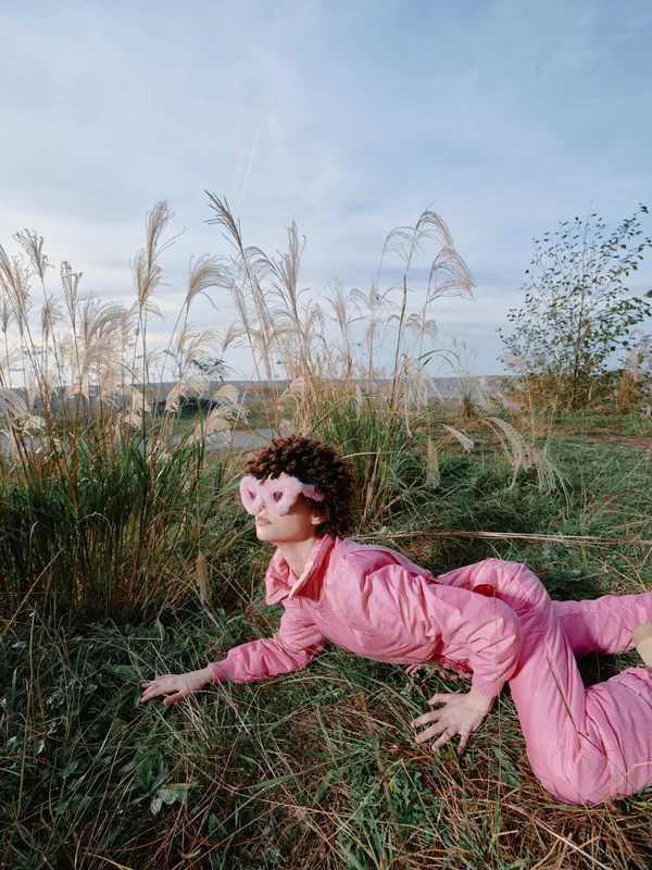 Outdoor scene with a person in a pink jumpsuit mid pose on a grassy field, arms stretched forward, a playful and free spirit captured beneath a soft cloudy sky.