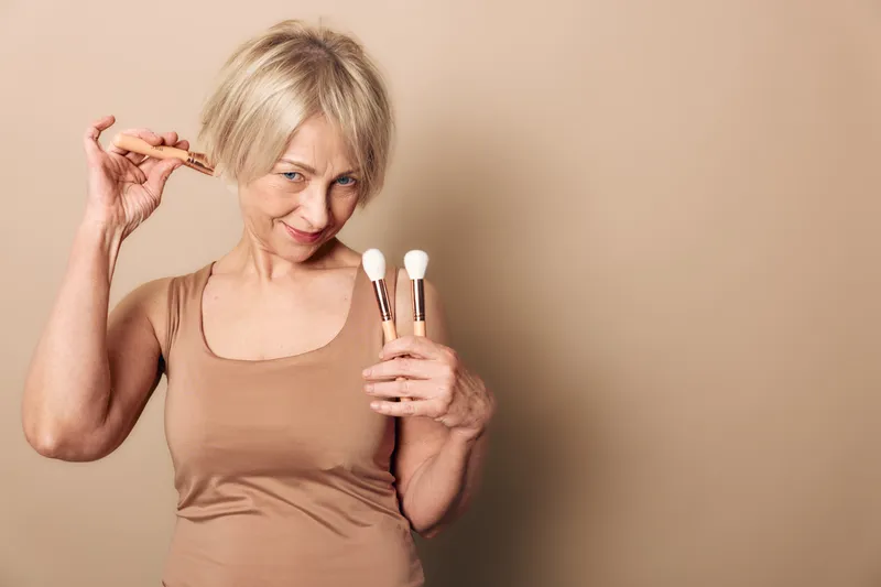 Smiling mature woman with short blonde hair holds makeup brushes and eyelash curler in beige top against neutral background. Concept for beauty tools, skincare routine, and self-care habits.
