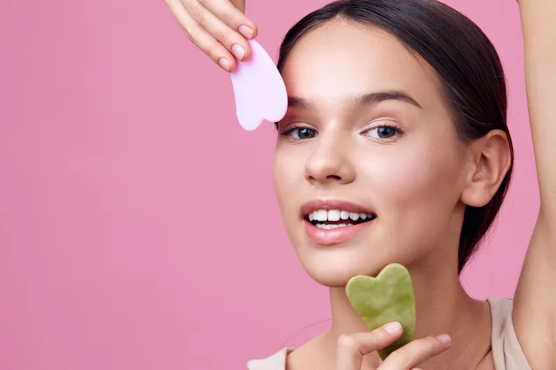 Young woman practicing skincare using gua sha tools on her smooth skin, showcasing relaxation and beauty against a pastel pink background