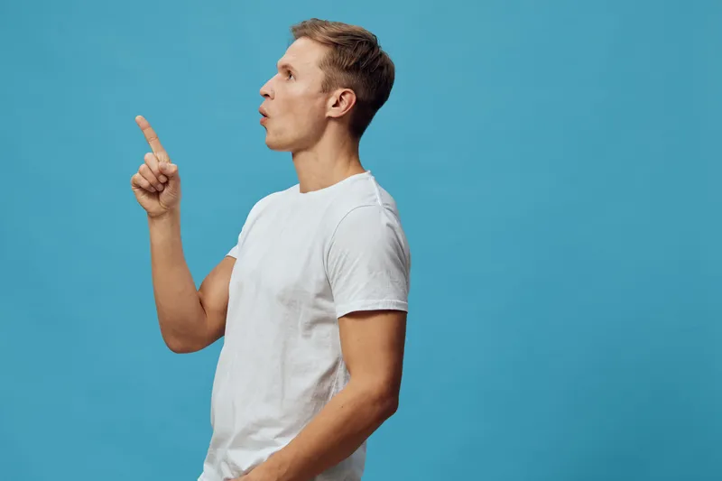Thoughtful young man with short hair wearing a plain white T-shirt standing isolated against a bright blue background. He is looking to the side, raising his finger, appearing to explain or emphasize 