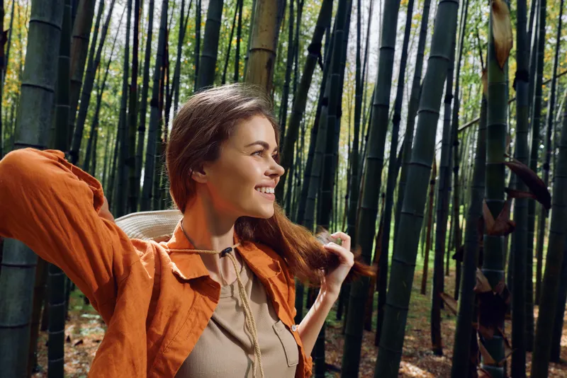 Woman in orange jacket smiling in a bamboo forest, portrait of happy traveler enjoying nature outdoors with sunlight filtering through tall bamboo stalks and peaceful green atmosphere.