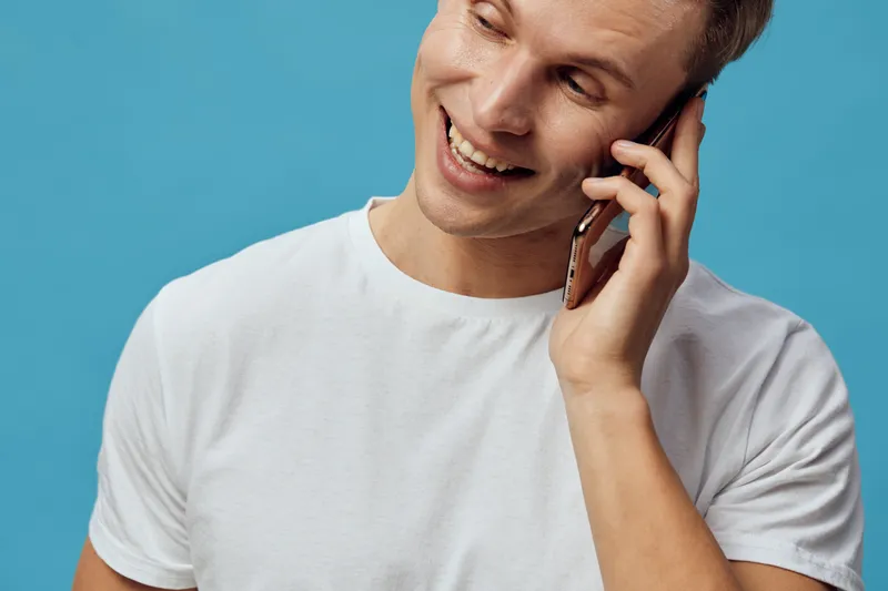smiling young caucasian man with smartphone in white t-shirt talking on phone isolated on blue background digital nomad concept communication technology