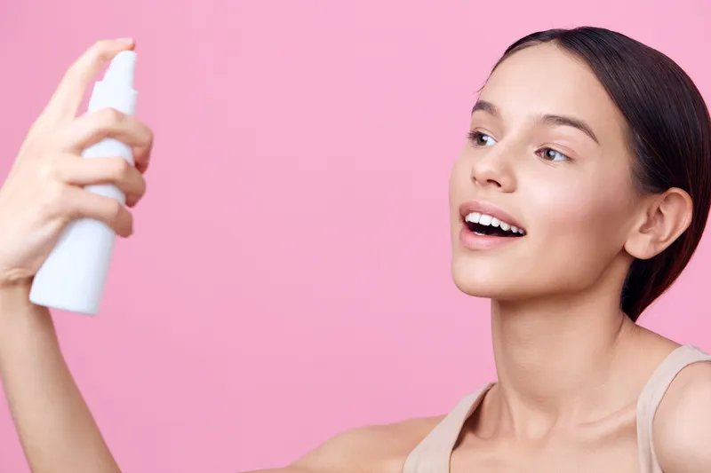 Young woman using a facial spray on a pink background, promoting skincare, beauty, and self care with an expression of joy and freshness