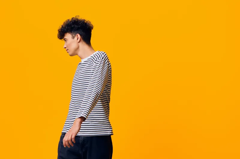 young man with curly hair wearing striped shirt standing sideways on vibrant yellow background, casual style and relaxed mood