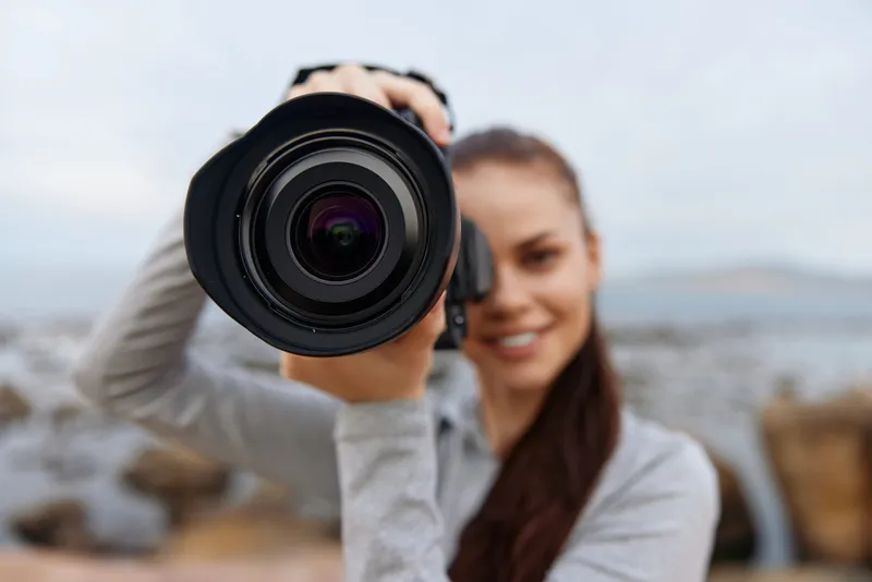 Young woman with long hair holds a camera on a rocky beach, smiling confidently in a casual outfit, capturing a moment in nature with a soft color palette
