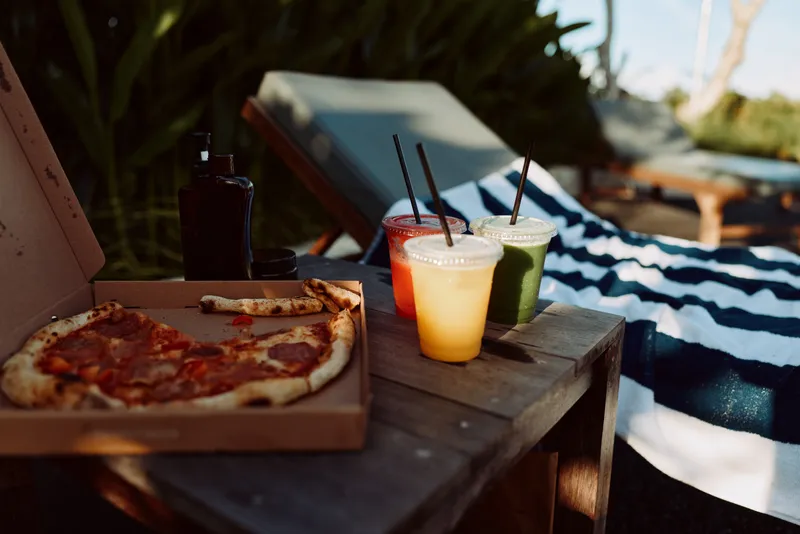 Casual outdoor picnic scene with a pizza on a wooden box, three colorful beverages on a table, striped blanket, lounge chairs, and bright sunlight creating a relaxed summer mood.