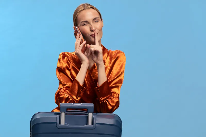 Blonde woman in orange blouse makes shushing gesture while holding dark blue suitcase on blue background. Concept for travel secrecy, quiet, and vacation preparation with studio lighting.