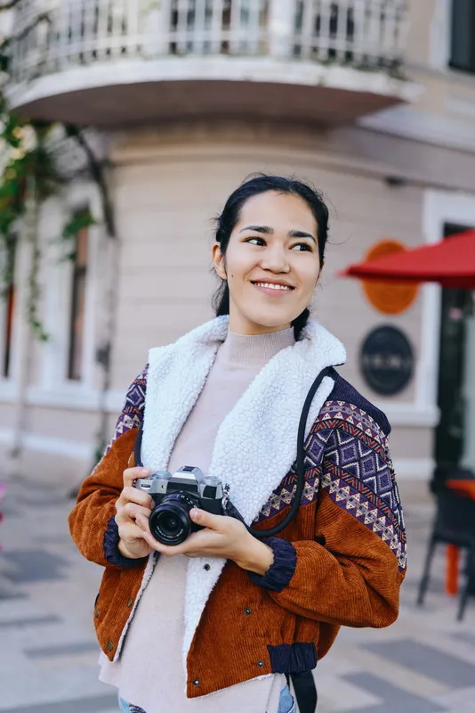 Street scene with a smiling woman holding a camera, stylish layered jacket, and fluffy scarf, capturing a casual urban moment outdoors, modeling lifestyle and travel vibes.