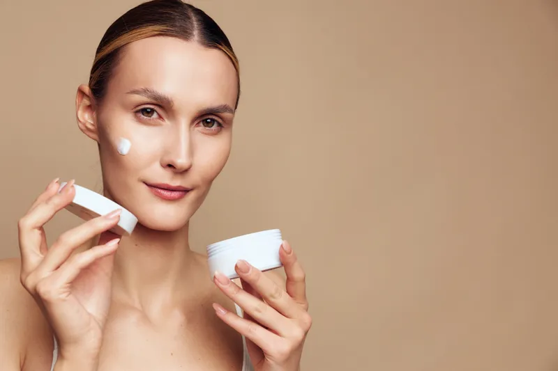 Young woman applying face cream, holding jar and lid, skincare routine, natural beauty, smooth skin, neutral background, soft lighting, close up portrait