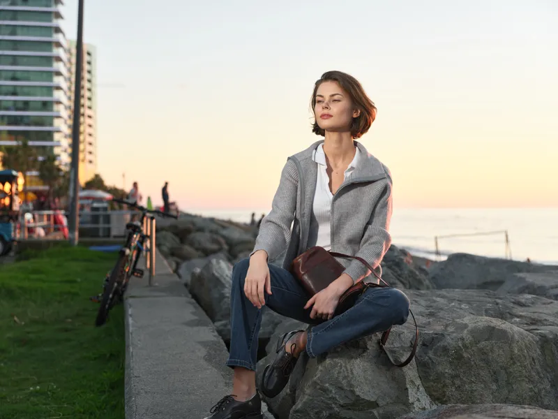 A stylish woman sits on a rocky seawall at sunset, dressed in a light jacket and jeans. She gazes toward the horizon with a calm, confident expression, creating a serene, urban coastal mood.