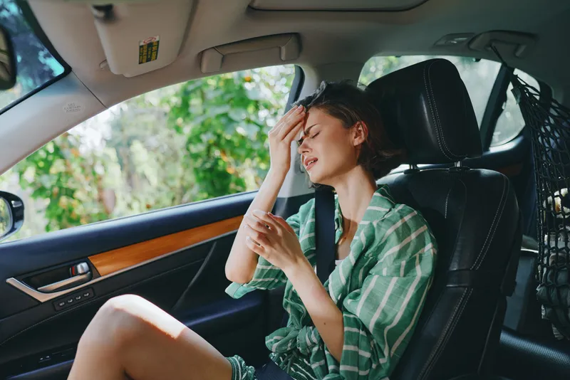 Young woman sits in the driver seat, hand on her forehead in a worried pose, casual green plaid shirt, daytime car interior.