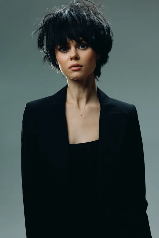 young woman with short black hair wearing black jacket posing against dark background