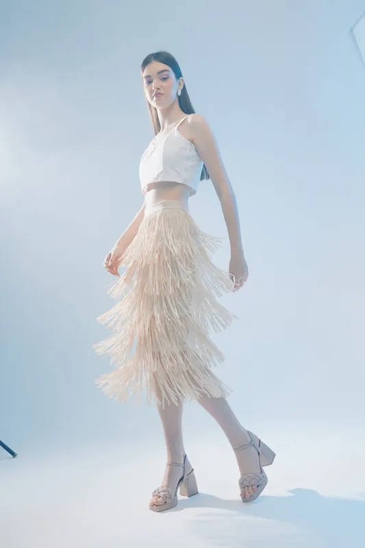 Young woman in a white crop top and fringed skirt poses in a studio with soft lighting. Concept of woman beauty fashion, elegance, and modern style.