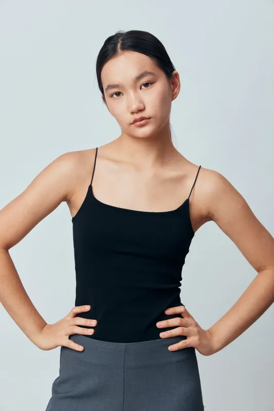 Young woman posing with hands on hips wearing black sleeveless top and gray pants, looking confidently at camera with neutral expression against plain background.