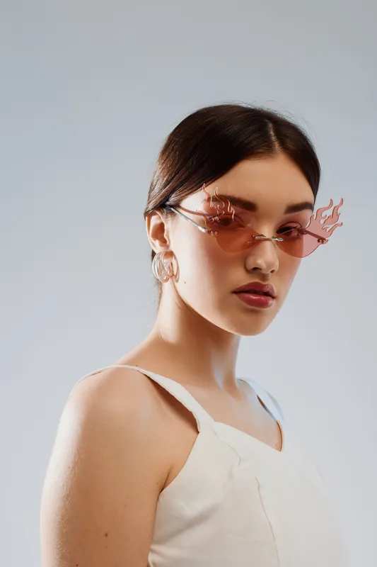 Young woman with dark hair wearing stylish flame-shaped sunglasses and white top poses against a plain background. Concept of woman beauty fashion and modern accessories.