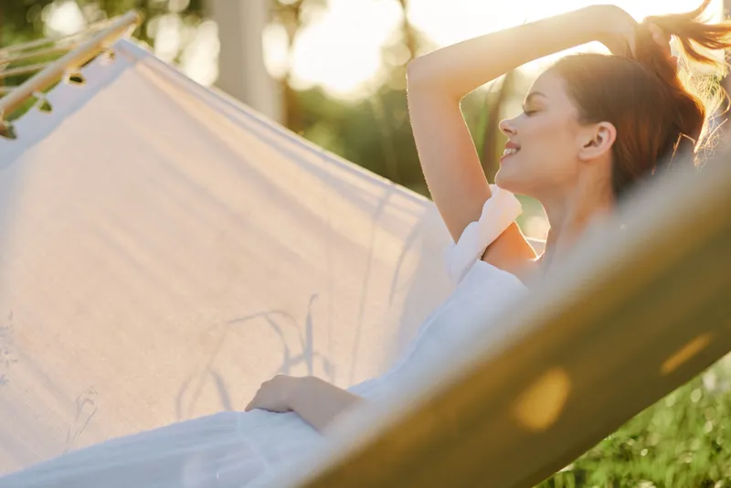 Relaxed young woman enjoying a sunny day in a hammock, wearing a light dress with a serene expression amidst green nature, ideal for summer and wellness themes