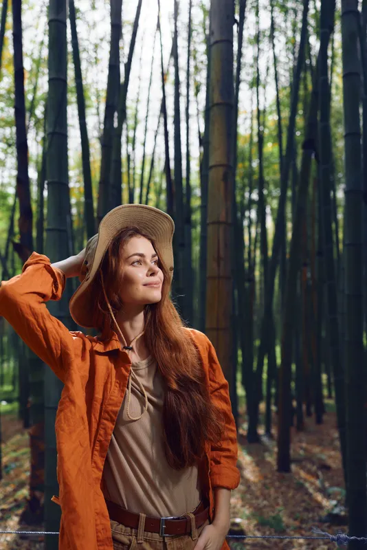 Woman with redhair and hat smiles in a bamboo forest, wearing orange jacket and casual outfit. Outdoor portrait of a young explorer enjoying sunlight, nature and peaceful grove.