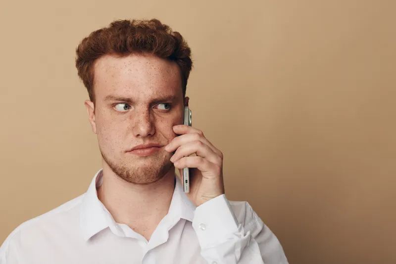 Young man with red hair and freckles wearing white shirt looks suspicious while talking on smartphone. Neutral beige background. Concept of distrust, doubt, and private conversation.