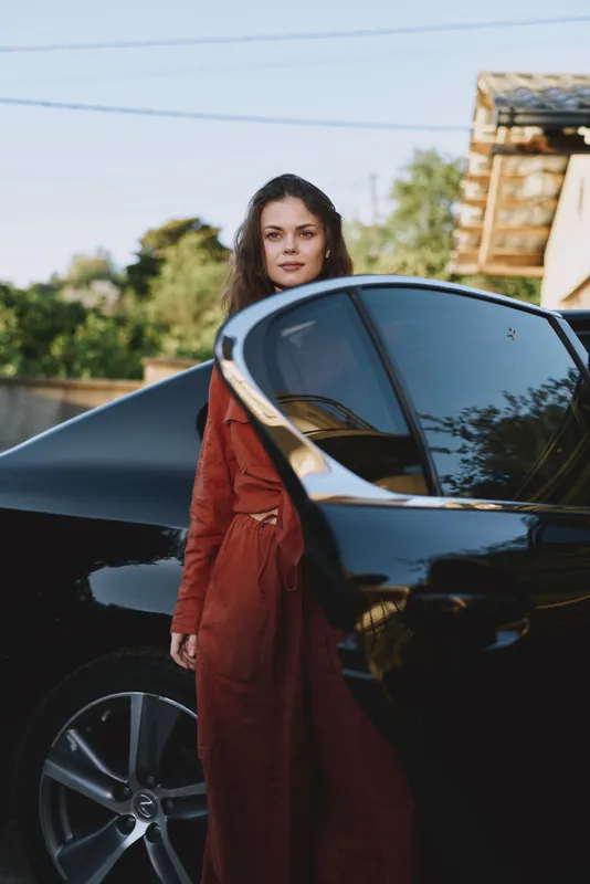 A stylish woman in a rust dress stands beside a sleek black car in a sunlit outdoor setting. The image captures confident, casual lifestyle moments and fashion inspiration for everyday chic