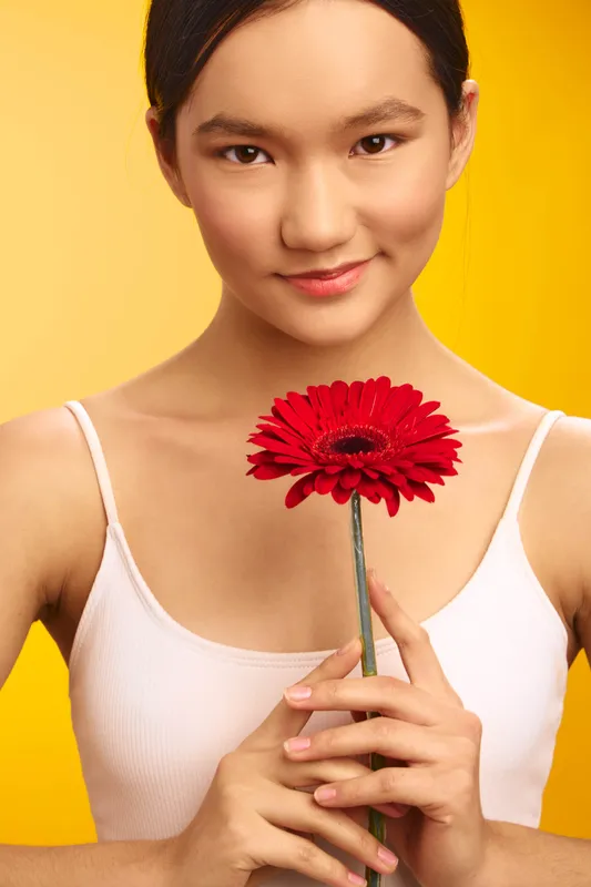Asian woman holds a red flower against a bright yellow backdrop, calm expression and soft smile creating warmth and elegance in a minimal studio portrait.