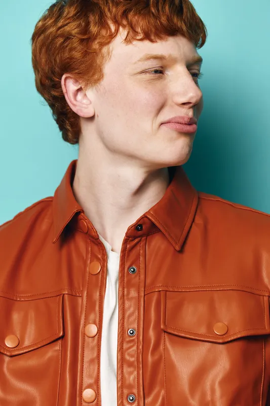 Young man with red hair wearing brown leather jacket and white shirt posing in profile against plain turquoise background with casual expression and natural light.