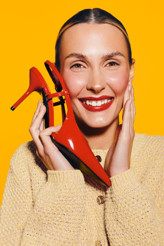 Smiling woman holds red high heel shoe near face on yellow background. Fashion concept with bright colors, beauty, and stylish footwear. Happy model in cozy sweater poses indoors.