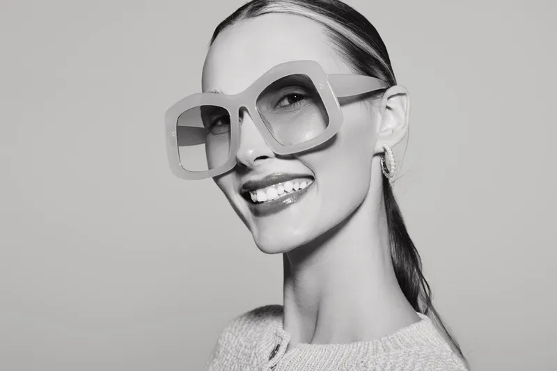 Slim white woman with pale skin wears large square glasses and smiles confidently on light grey background. Concept of fashion, eyewear, and beauty.