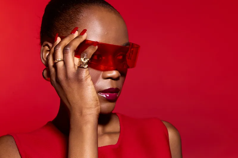 Stylish black woman with dark skin wearing red sleeveless top and red transparent glasses poses against red background. Concept of fashion, beauty, elegance, and modern style.