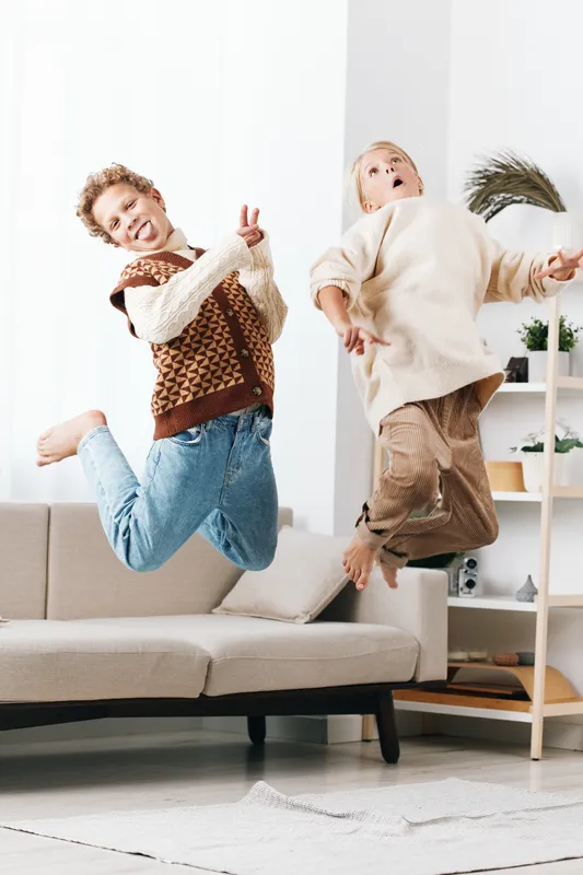 Two joyful friends jump on a sofa in a bright living room, wearing cozy sweaters and jeans, capturing energy, playfulness, and a spontaneous, carefree moment.
