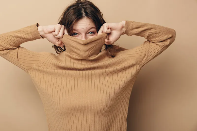 Young woman in brown knitted sweater pulls collar over face, smiling with playful eyes on beige background. Concept of cozy fashion, autumn style, and casual comfort indoors.