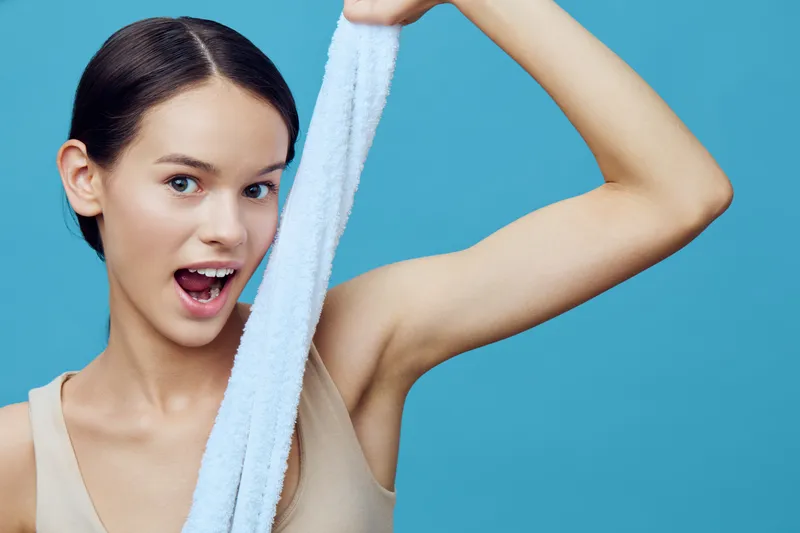 Young woman with a cheerful expression holding a towel against a light blue background, showcasing fitness and healthy lifestyle concepts