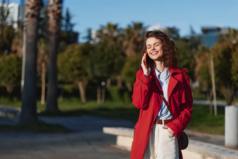 Young woman with curly hair wearing a red coat smiles happily while talking on her phone outdoors on a sunny day in a park setting.