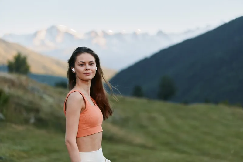 A cheerful young woman stands on a grassy hillside with distant mountains in the background. She wears a peach top, smiles softly, and enjoys warm natural light in an outdoor portrait.