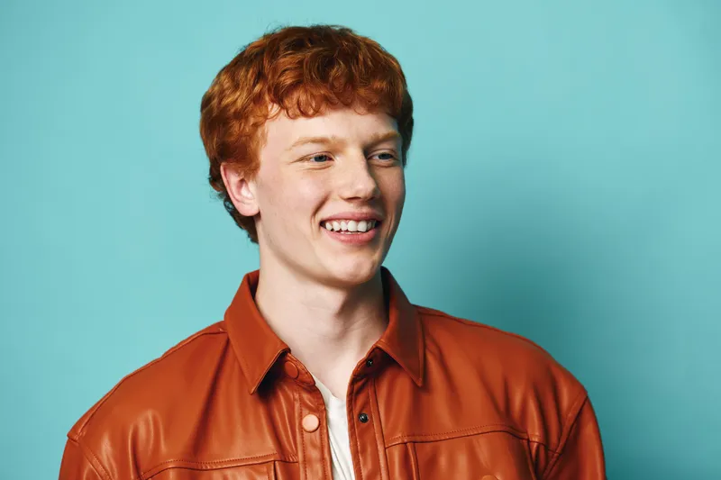 Young man with red hair and smiling face wearing orange shirt stands against blue background in studio. Casual portrait with happy expression and natural lighting for positive mood.