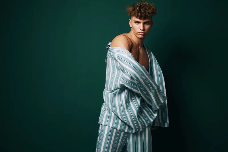 Young man with curly hair wearing striped pajamas posing against dark green background, fashion portrait with casual style and confident expression in studio lighting.