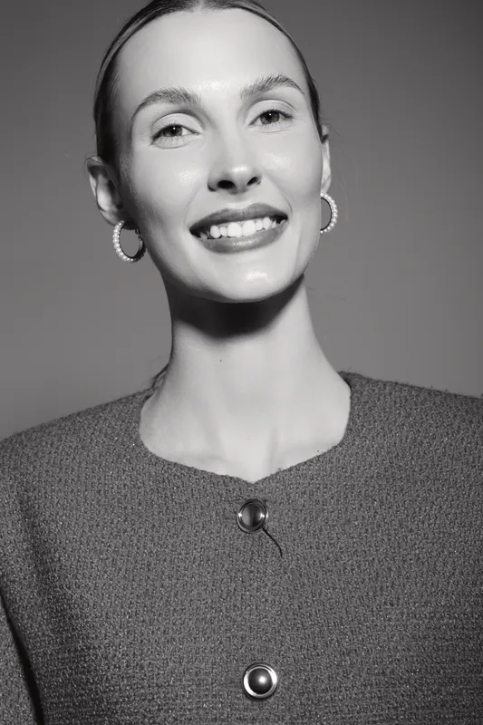 Smiling Caucasian woman with fair skin wearing textured jacket and hoop earrings posing on grey background, portrait with soft lighting.