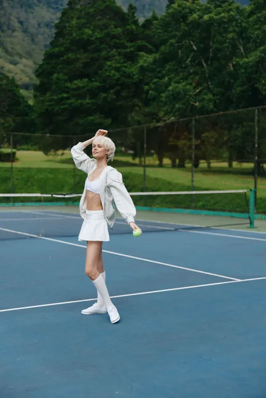 Young athletic woman celebrating a tennis victory in a stylish white outfit on a blue court surrounded by greenery, expressing joy and confidence