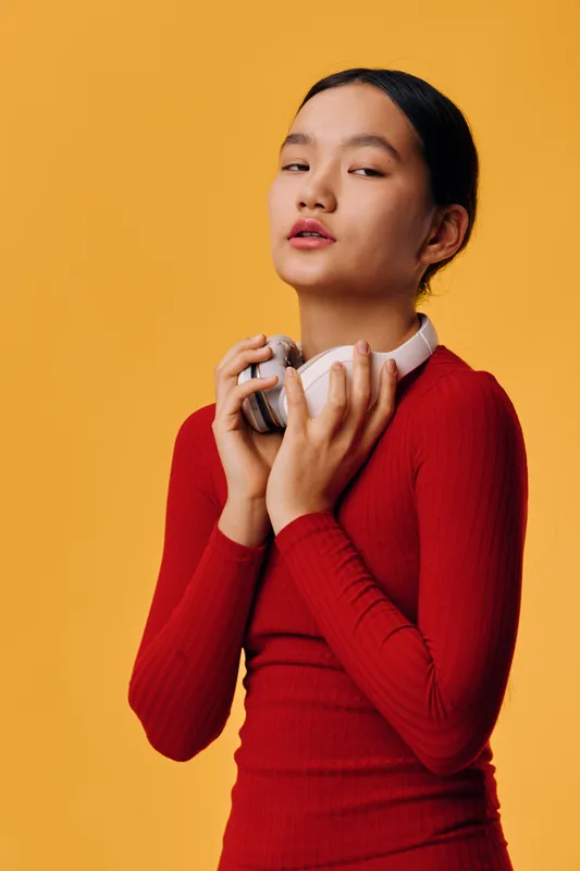 Young woman in red sweater holding white headphones close to chest with eyes half closed against orange background. Portrait of serene girl with dark hair in minimalist studio setting.