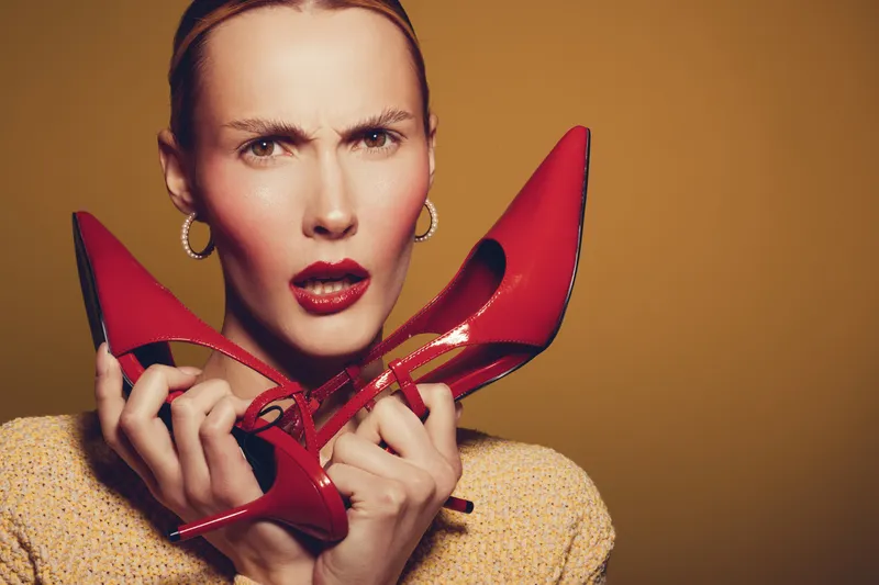 Confused woman with red lipstick holds red high heel shoes near face on yellow background. Concept of fashion dilemma, style choice, and female expression of uncertainty.