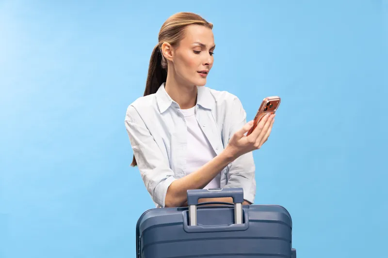 Young woman with suitcase looks at smartphone, dressed casually, isolated on blue background. Concept of travel planning, communication, and modern lifestyle.