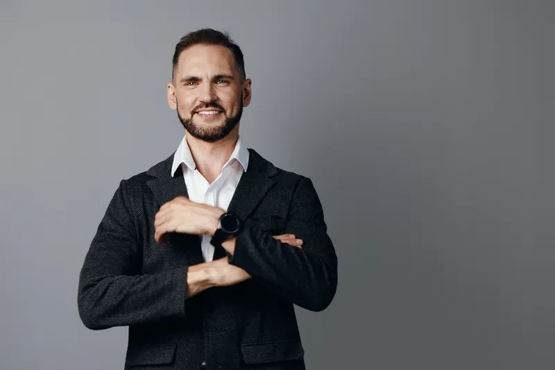 A confident business professional stands against a clean colored backdrop, arms crossed, smiling subtly to convey approachability, focus, and leadership in a modern work environment.