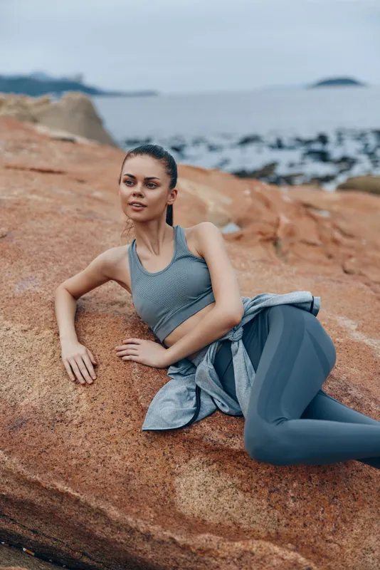 Young woman in activewear sitting on rocks by the sea, exuding a calm and determined expression, embodying fitness and healthy lifestyle concepts in a coastal setting