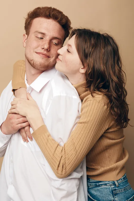 Young couple embraces tenderly, woman hugging man from behind with closed eyes. Warm beige background, casual clothing. Concept of love, affection, intimacy, and relationship.