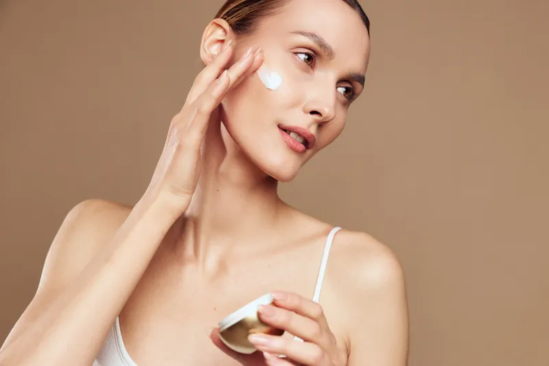 Young woman applies face cream on cheek with fingers holding open jar skincare beauty routine isolated on beige background natural skin care concept.