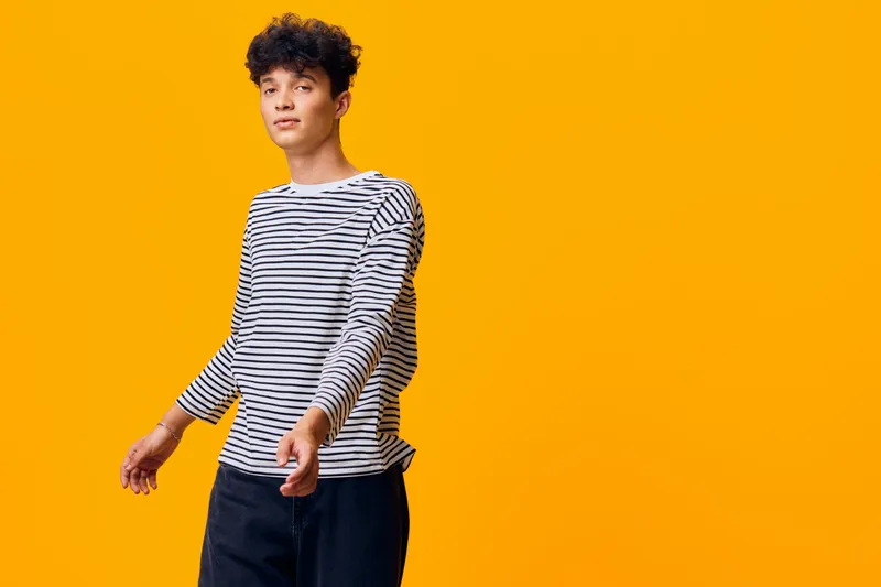 young man wearing striped shirt poses confidently against bright orange background, casual style with curly hair and relaxed expression in modern studio setting